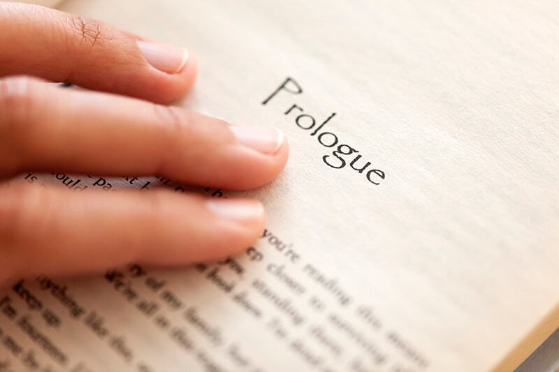 Close up image of a book page with the text "prologue" at the top. There is a hand touching the page right below the font.
