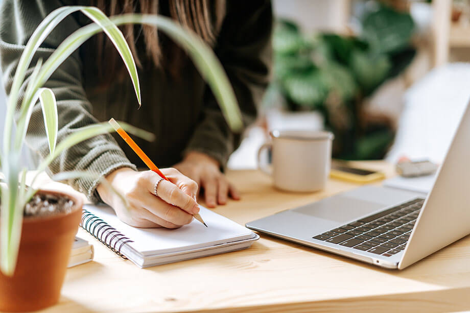 Woman writes in notebook in front of laptop while surrounded by desk plants