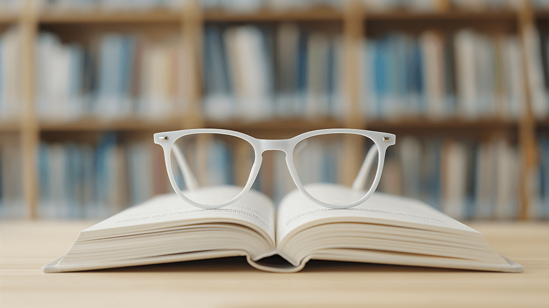 Open book with white glasses resting on the pages on a wooden table in front of a bookshelf in a library